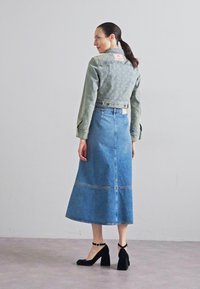 Jeans jakna s suptilnom uzorkovanom teksturom, uparena s plavom dugom trakom od traperica i crnim cipelama na blok povišenoj peti, prikazana s leđa.
