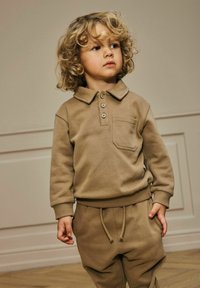 Niño pequeño con cabello rubio rizado, vistiendo una camiseta polo de manga larga marrón y pantalones con cordón a juego, de pie en el interior sobre un suelo de madera.
