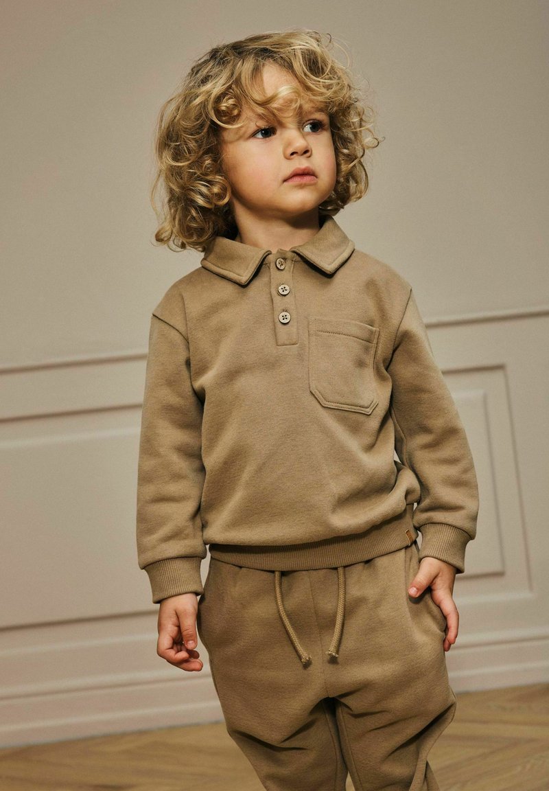 Niño pequeño con cabello rubio rizado, vistiendo una camiseta polo de manga larga marrón y pantalones con cordón a juego, de pie en el interior sobre un suelo de madera.