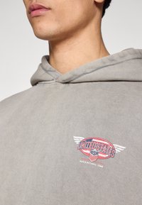 Γκρι φούτερ με κουκούλα από απαλό ύφασμα, με κυκλικό σχέδιο πο�υ απεικονίζει "UNITED STATES" και φτερά σε κόκκινο, λευκό και μπλε.