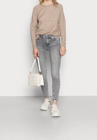Beige stickad tröja, grå slim fit jeans, vit vävd handväska och lätta sneakers med accentdetaljer. Enkelt, avslappnat outfit mot en neutral bakgrund.