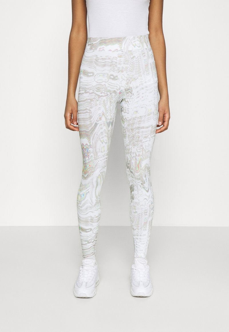 Kvinna i högmidjade vita och grå mönstrade leggings med vita sneakers, stående mot en helt vit bakgrund.