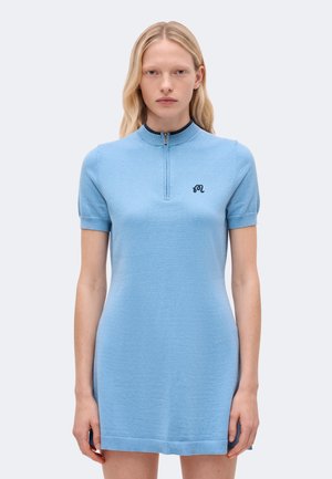 Une femme porte une robe en maille bleu clair à manches courtes, dotée d’une fermeture éclair sur un quart de la longueur et d’un petit logo sur la poitrine, devant un fond uni.