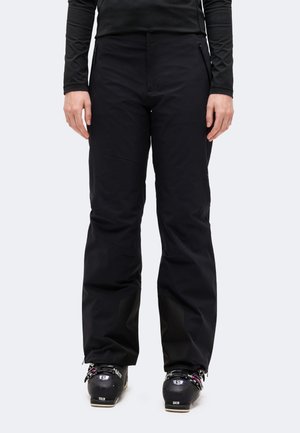 Une personne présente un pantalon noir avec des poches zippées et des chaussures de ski, sur un fond uni clair.