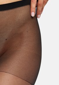 Pantys negros transparentes con un acabado texturizado y una cinturilla ancha y elástica, con una mano señalando la superficie del material.
