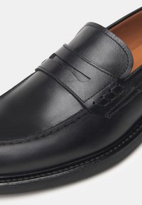 Mocasines de cuero negro con un acabado suave, una correa decorativa, costuras en contraste y una punta redondeada. El interior es de color marrón claro.