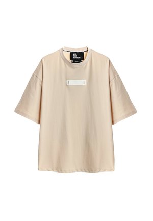 Camiseta beige oversize de manga corta con cuello redondo y un pequeño parche rectangular en el pecho.