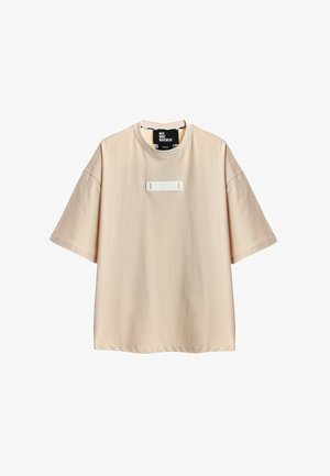 Camiseta beige oversize de manga corta con cuello redondo y un pequeño parche rectangular en el pecho.