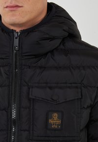 Giubbotto nero in stile puffer con chiusura a zip, grande tasca sul petto e toppa con logo. Tessuto trapuntato; dispone di un cappuccio e colletto a coste.