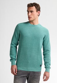 Turquoise gebreid sweater met ribbeltextuur, ronde hals en lange mouwen. Heeft een contrasterende onderrand en manchetten, aangevuld met subtiele branding.