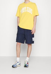 Žluté bavlněné tričko s bílým nápisem "TOMMY JEANS" v obloukovém písmu, spárované s námořnickými cargo šortkami a multicolor teniskami s vzorovaným designem.