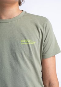 Olijfgroene katoenen t-shirt met ronde hals, korte mouwen en bedrukte tekst in felgeel aan de linkerzijde. Glad textuur.