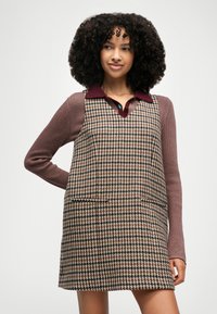 Klänning med brun- och svart hundtandsmönster och sidofickor, bärs över en vinröd skjorta med krage och lila ribbade ärmar.