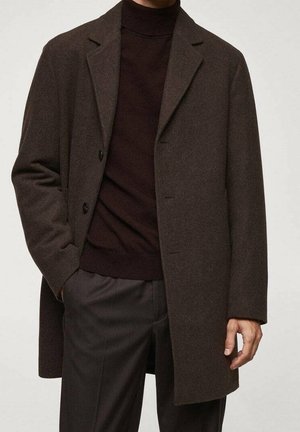 Homme portant un manteau en laine brun foncé sur un pull à col roulé noir et un pantalon foncé, main partiellement dans la poche du manteau, debout devant un fond uni.