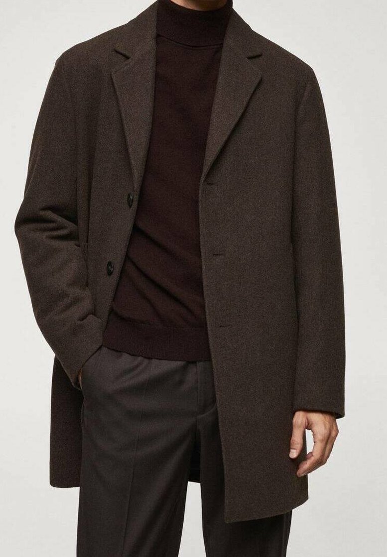 Homme portant un manteau en laine brun foncé sur un pull à col roulé noir et un pantalon foncé, main partiellement dans la poche du manteau, debout devant un fond uni.