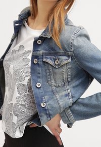Lysblå denimjakke med sølvknapper, to brystlommer og en let slidt finish. Bæres over en sort-hvid blomstret t-shirt med grafik.