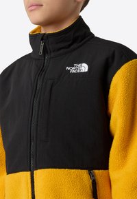 Μαύρο και κίτρινο μπουφάν από φλις με ψηλό γιακά, φερμουάρ μπροστά και τσέπη στο στήθος με φερμουάρ. Διαθέτει αντίθετες υφές και κεντημένο λογότυπο.