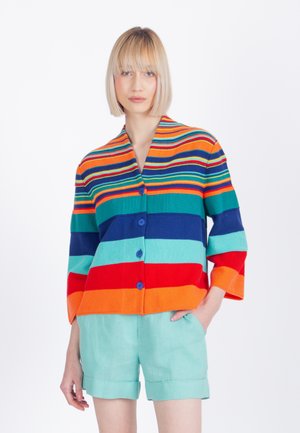 Mladá žena s blond bobom, oblečená v farebnom pruhovanom kardigáne na gombíky a svetlo tyrkysových šortkách, stojaca pred bielym pozadím.