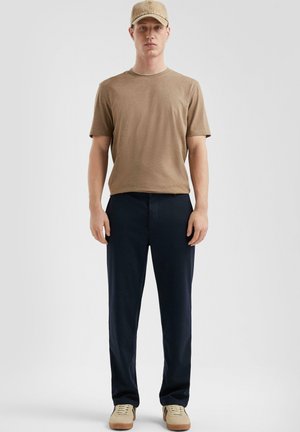 Tan korte mouwen t-shirt, donkerblauwe broek, beige sneakers en een beige pet. Eenvoudig ontwerp met een ontspannen pasvorm en geen zichtbare patronen.