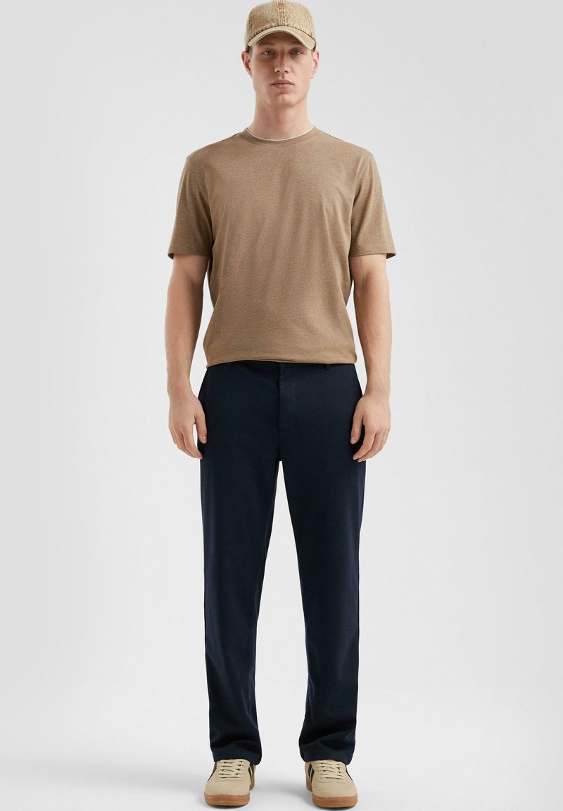 Tan korte mouwen t-shirt, donkerblauwe broek, beige sneakers en een beige pet. Eenvoudig ontwerp met een ontspannen pasvorm en geen zichtbare patronen.