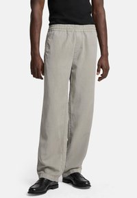 Lichtgrijze, loszittende broek met een elastische tailleband en zijzakken, gecombineerd met zwarte schoenen. Gladde stofstructuur.