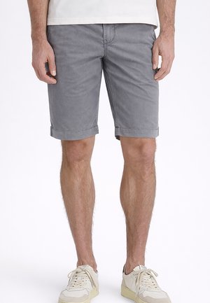 Mann, der aufgerollte graue Shorts, weiße Sneakers und ein weißes Hemd trägt, steht vor einem schlichten weißen Hintergrund.