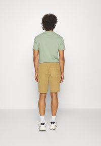 T-shirt vert à manches courtes associé à un short beige clair. Le short a une poche arrière, les deux vêtements ont une coupe décontractée. Baskets blanches avec des accents noirs.