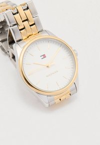 Hodinky z nerezové oceli ve stříbrné a zlaté barvě s bílým ciferníkem, minimalistickými hodinovými značkami a akcentem loga. Náramek ve stylu řetízku.