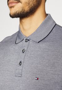 Šedé polo tričko s texturovaným povrchem, s pruhovaným límcem, třemi knoflíky a malým logem na hrudi.