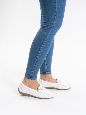 Person wearing slim blue jeans and white slip-on loafers with gold buckle details, standing on a white background.
