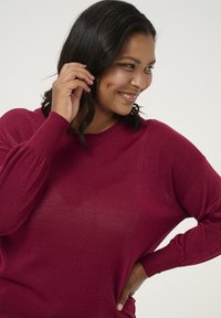 Sweater en tricot bordeaux à manches longues, avec une coupe décontractée, des poignets côtelés et une texture légèrement brillante.