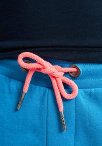Modré tepláky mají tkanou strukturu, růžovou šňůrku uvázanou do mašle a kovovou kroužkovou ozdobu na pase.