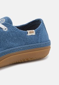 Sapatilha de lona azul com atacadores brancos, sola de borracha bege e etiqueta da marca Vans. Apresenta um material texturizado e detalhes costurados ao longo da sola.