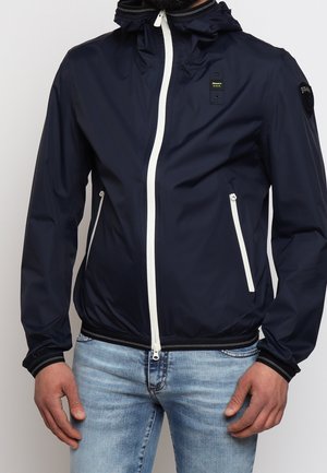 Mand iført en marineblå jakke med hvide lynlåse og lommedetaljer, kombineret med lyseblå jeans, stående foran en ensfarvet baggrund.