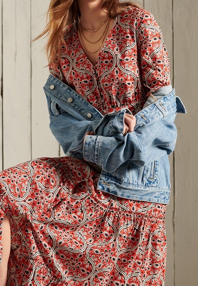 Femme portant une robe rouge à motifs avec une veste en jean bleue posée sur ses épaules et des colliers dorés superposés.