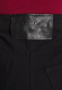 Pantaloni in denim nero con un'etichetta in pelle texturizzata con la scritta "PATRIZIA PEPE" in rilievo, esposti su uno sfondo rosso.