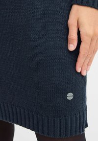 Marinblå stickad klänning med ribbad nederkant och manschetter, prydd med en liten cirkulär metall-logotypdetalj nära botten, slät textur och åtsittande silhuett.