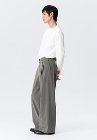 Homme en chemise blanche à manches longues et pantalon large à rayures grises, se tenant de profil avec les mains croisées derrière le dos sur fond blanc.