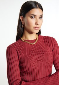 Roter, ribbeliger Pullover mit strukturiertem Muster und rundem Halsausschnitt. Accessoires: eine dicke goldene Kette und florale Ohrhänger.