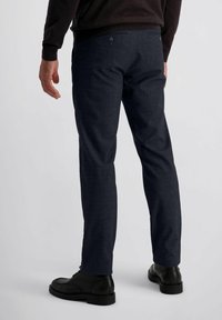 Man in een donkere geruite slim-fit broek en zwarte leren veterschoenen, staande tegen een effen lichte achtergrond.