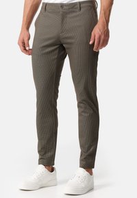 Pantaloni a quadri marroni con vestibilità slim, dotati di chiusura a bottone, tasche laterali e una texture di tessuto morbido. Indossati con sneakers bianche.
