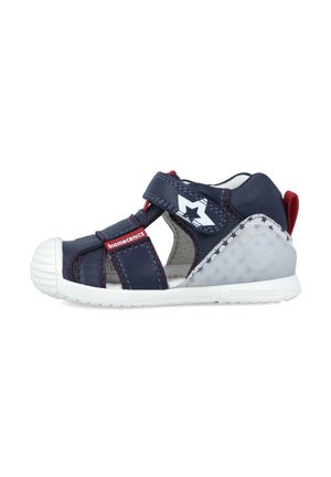 Sanda blue navy con acentos rojos, correa ajustable y suela de goma blanca. Presenta un diseño de estrellas en el lado y perforaciones transpirable.