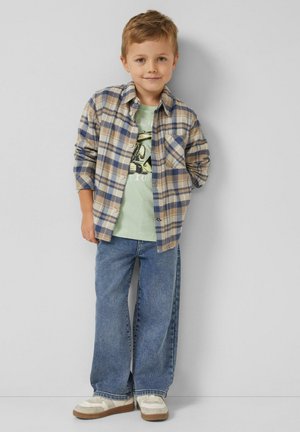 Junge in einem beige-blauen karierten Flanellhemd über einem hellgrünen T-Shirt, trägt lockere blaue Jeans und weiße Turnschuhe mit braunen Sohlen.