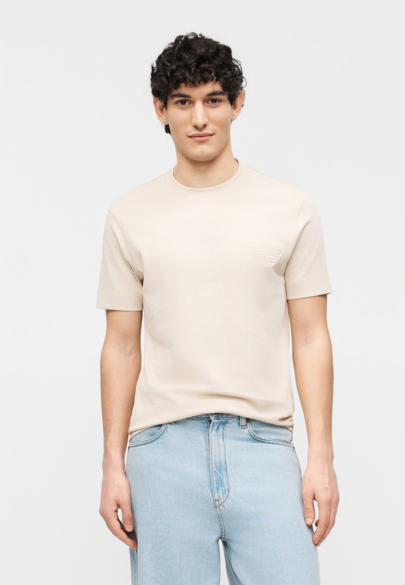 Νεαρός άνδρας με κοντά κατσαρά μαλλιά φοράει μπεζ κοντομάνικο μπλουζάκι και ανοιχτόχρωμα μπλε τζιν μπροστά σε απλό λευκό φόντο.