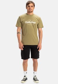 Oliven grøn t-shirt med Quiksilver-logo, parret med sorte cargo shorts. Modellen har hvide ankelstrømper og sorte sneakers på. Simpelt, afslappet design.