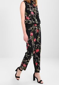 Svart blommig overall med röda och vita blommönster, ärmlös design, lös passform och smalnade byxor. Bärs med svarta högklackade sandaler.