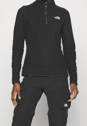 Osoba ubrana w czarną polarową kurtkę The North Face z półzamkiem oraz czarne spodnie cargo z klapkami, oba z logo The North Face.