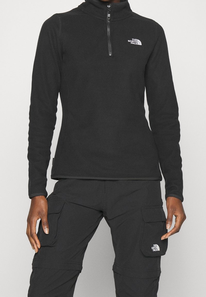Personne portant une veste polaire noire North Face avec fermeture éclair mi-longue et un pantalon cargo noir avec des poches à rabat, les deux avec le logo The North Face.