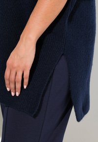 Mörkblå textured stickad tunika med sidofack, kombinerad med mörka byxor. Närbild av en hand som vilar på plaggkantens kant.