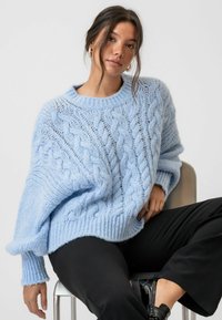 Ljusblå chunky stickad tröja med ett kabelmönster och ribbade ärmslut och nederkant; kombinerad med svarta vida byxor och skor.